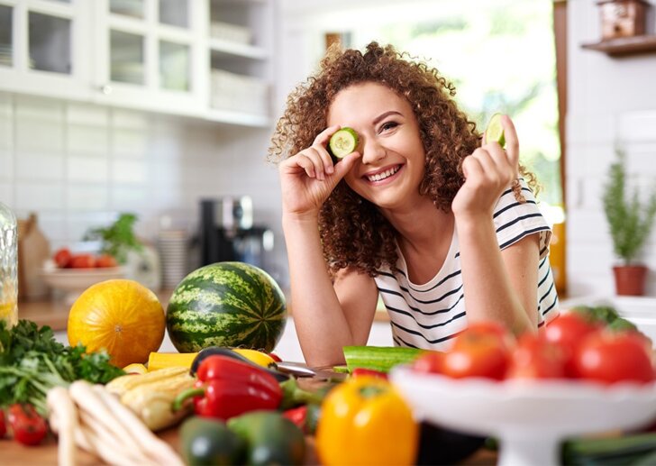 Eine Jugendliche steht in der Küche, Obst und Gemüse liegen vor ihr und hält sich eine Scheibe Gurke vor das rechte Auge  | © iStock.com/gpointstudio
